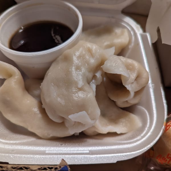 Steamed dumplings and a cup of dark dipping sauce in a white takeout container.