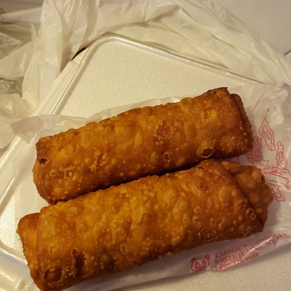 Two crispy, golden-brown egg rolls resting on white paper inside a styrofoam takeout container.