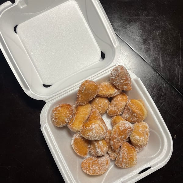 A white styrofoam takeout container filled with golden-brown, sugar-dusted donut holes on a dark surface.